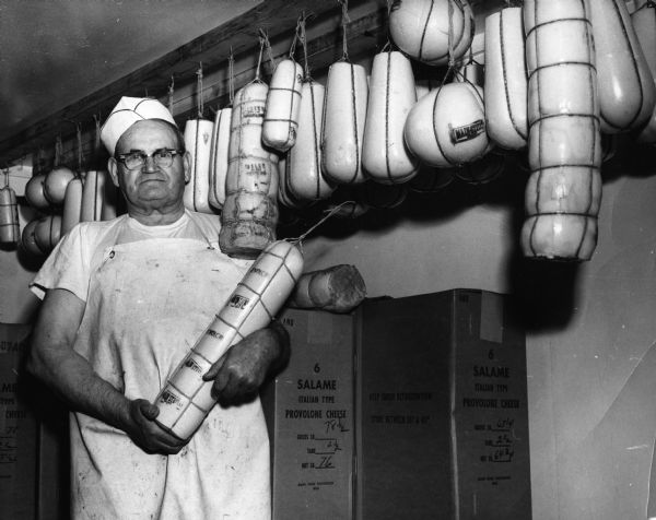 Mike Cotch standing holding a provoloni with other shapes of provolone cheese including salomini, salame, and madget hanging from the ceiling at the Burnett Dairy Co-Op.