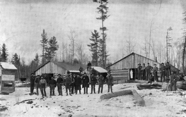 Lumber company workers pose in front of buildings at the Moore & Galloway Lumber Company camp, about a quarter of a mile east of Pike Lake. The man in a white apron standing in a doorway is Ole P. Dobbe of Alban, who was the cook. The building in the center was probably the woods boss's office, and the building to its left is a barn. The small building at the extreme left may be an outhouse.