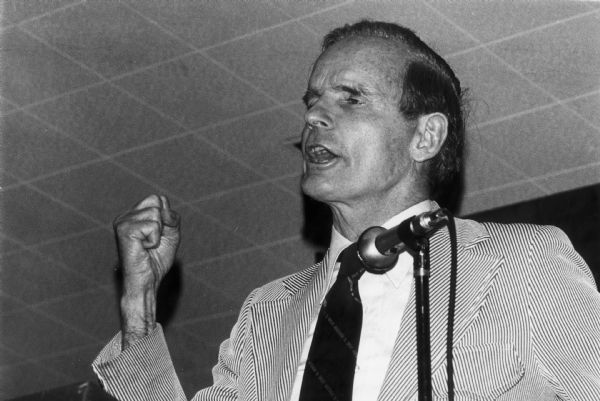 William Proxmire gives a speech wearing a striped suit and raising his fist.