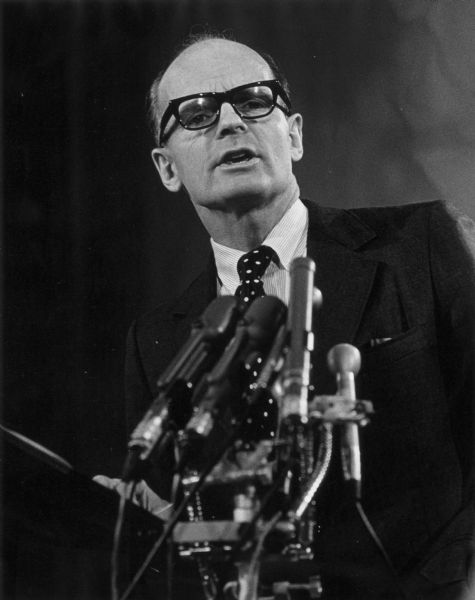 Senator William Proxmire gives a speech in front of many microphones, wearing eyeglasses.