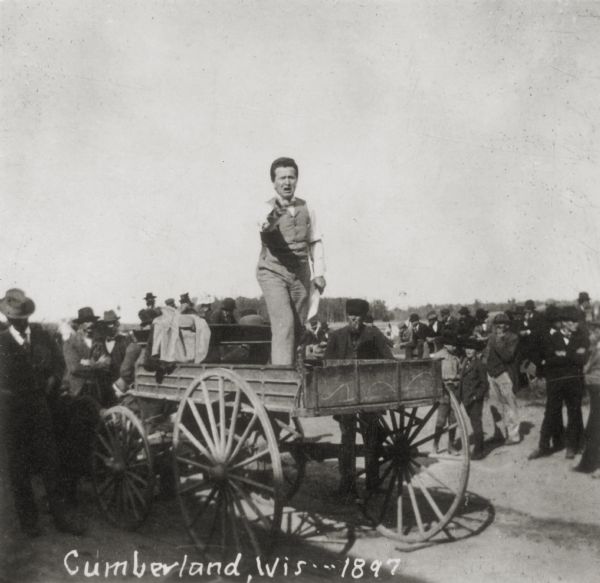 Robert M. La Follette, Sr., speaking to a crowd from back of a wagon.  This image is one of a series of views of his appearance at a fair in Cumberland, Wisconsin, in 1897. During this time La Follette made a series of county fair speeches primarily aimed against machine politics and advocating a secret ballot primary election. Part of La Follette's success in eventually winning the Wisconsin gubernatorial election in 1900 was a result of  the many reform speeches he had delivered to local audiences during the previous years. La Follette's vigorous speaking style had much to do with his eventual electoral success.