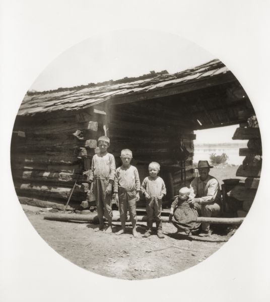 A man poses with his four children, who are lined up next to him from the tallest to the smallest, in front of a log shanty. The building is near the mouth of the Saline River, which can be seen in the background.