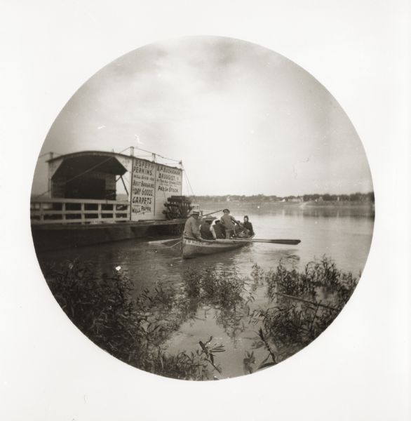A rowboat full of passengers floats alongside a ferry, which travels between Rabbit Hash, Kentucky, and Rising Sun, Indiana. On the side of the ferry are signs for Espey and Perkins, Dry Goods, and for B.F. Buchanan, Druggist.