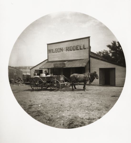 Exterior view of the Wilson Riddell general store, with a horse-drawn wagon parked in front. There is a post office and paint shop attached to the store. Several men are standing behind the wagon.