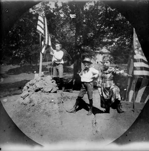 Fred Thwaites, Harry Turville, and Helen Turville celebrate the Fourth of July with American flags outdoors. There is a barn in the background.