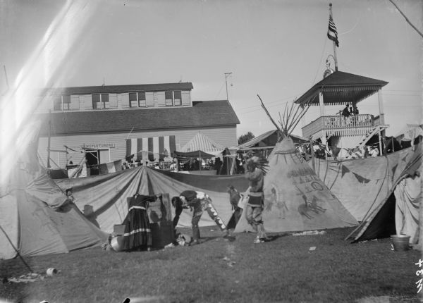 Native American Indians in traditional dress outside their teepees (tipis) at the Dodge County Fair. There is a large building in the background along with a viewing pavilion which has a large clock and flagpole on the roof.
