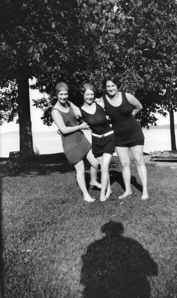 Mary Brandel, her mother, and a friend pose together wearing bathing suits. Trees and a lake are behind them, and the shadow of the photographer can be seen on the lawn.