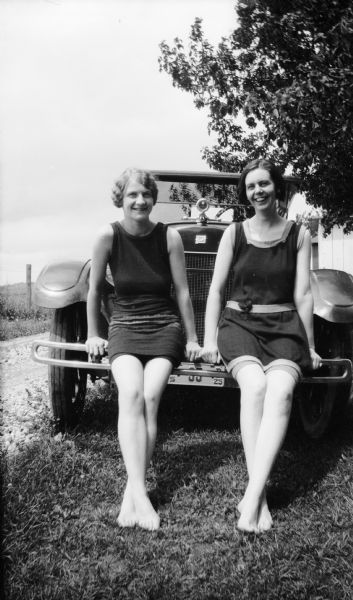Mary Brandel and a friend sit on the front of a car. They are wearing bathing suits. A road and fence are on the left.
