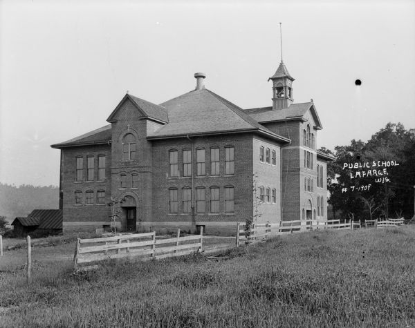Exterior view of the public school. A large brick building with a bell tower on the roof, arched entrances, and a wooden fence along the side.