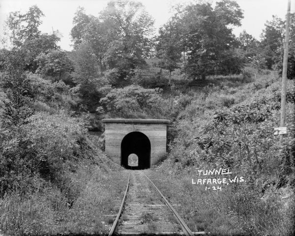 View from railroad tracks through covered railroad tunnel in hill. A sign on a telephone pole on the right reads: "Private Property / Thoroughfare / Keep Off." Carving above arch of train tunnel says, "AD 1900" (the D is backwards).