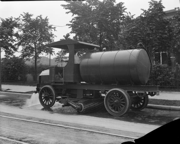 Side view of a man driving a street cleaning truck with water reservoir, sprayer, pump and brushes. Tracks are running down the street, perhaps for a cable car.