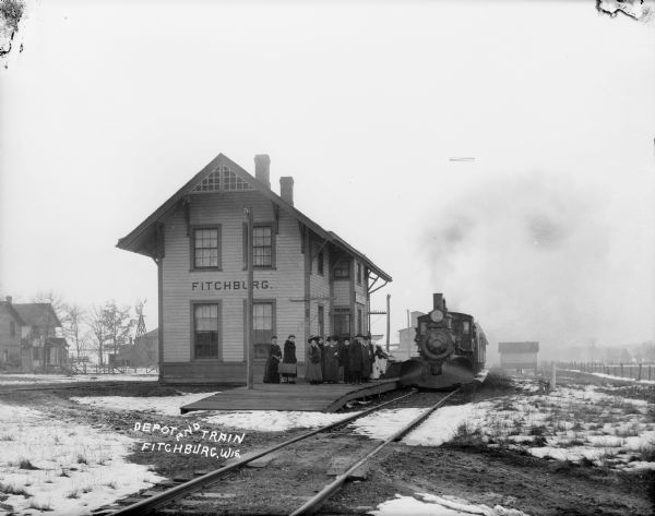 Winter scene of Fitchburg depot with train at platform, and a group of men and women waiting outdoors.