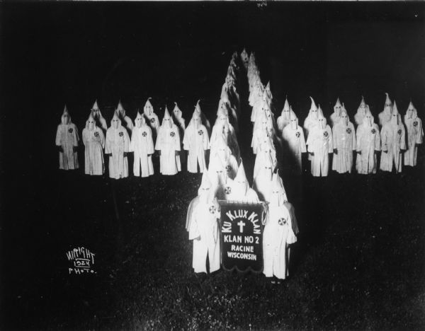 Elevated view of Ku Klux Klan (KKK) of Racine, posed outdoors in the form of a cross and holding a banner.