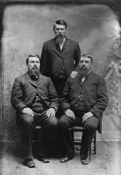 Studio portrait in front of a painted backdrop of the three Pond brothers, George F., James B., and Homer. They all have beards and moustaches. Two of the men are seated and one stands between them.