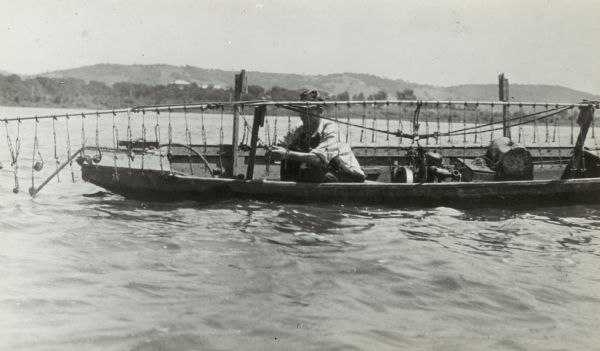 A clam fisher in a boat on the Mississippi River near Prairie du Chien. Attached to the long rod are many small hooks similar to grappling hooks. As the boat drifts along or is forced ahead by the motor, the rod is lowered and the hooks, trailing in the mud of the river bottom, hook into clam shells. In the picture the fisherman is removing his catch, the rod resting on wooden supports. In the background is the far shoreline.