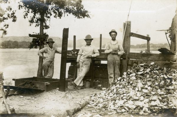Portrait of three men standing on the banks of a body of water next to a pile of clam shells and a wooden construction (possibly a dock).  The men are each wearing overalls and hats and are clam shell fishers and pearl hunters. The man on the left is identified as probably Rabbit Pyers.
