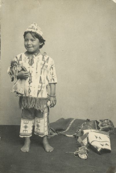 Studio portrait of Zintka Lanuni ("Lost Bird") Colby, a Lakota, as a young child wearing traditional Indian clothing of a fringed shirt, and pants. She is barefoot and is holding a doll under her right arm, and a ball in her left. She also wears a necklace, ring and bracelet, and other items, a blanket, and perhaps a moccasin are on the floor.