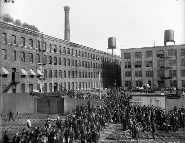 Elevated view of workers filing out of the gates of the McCormick Reaper Works at the end of a work day. The factory became part of the International Harvester Company in 1902. There are railroad tracks in front of the gates. Some of the workers carry lunch pails, and some have bicycles.