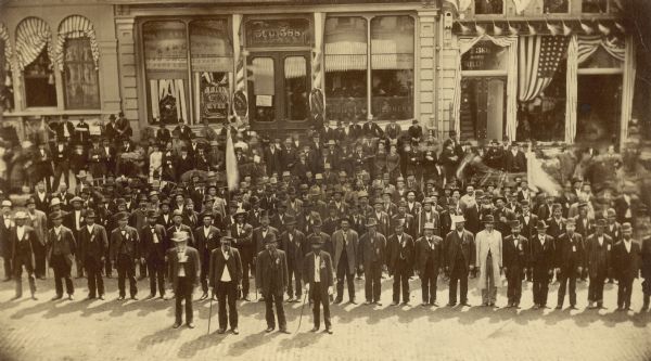 Elevated group portrait of the veterans of the First Wisconsin Calvary. They are standing in rows in the street. Horses with wagons are behind them, and a crowd is on the sidewalk in front of storefronts decorated with bunting and flags.
