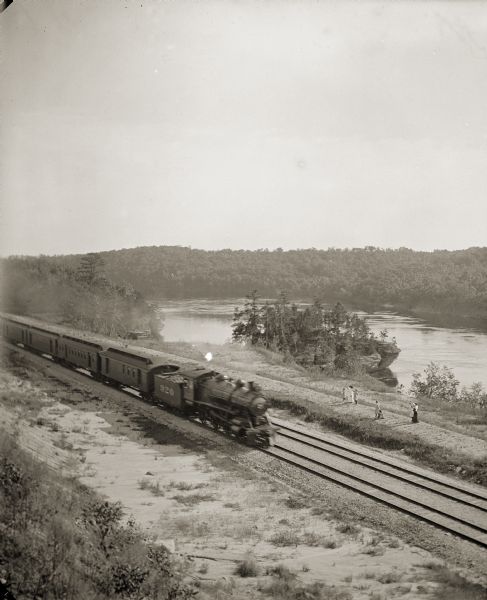 Elevated view of a train running parallel to the river, past the Ink Stand. Several people are standing along the tracks and waving.