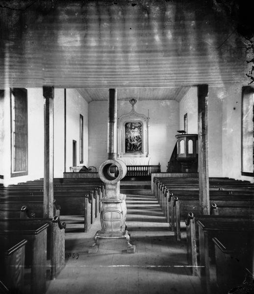 View of the interior of a small Lutheran church. A wood-burning stove is in the aisle between the pews, and a painting is hanging above the altar.