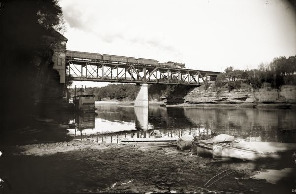 View of Kilbourn bridge with railroad train. Steamboat and rowboats at edge of river.