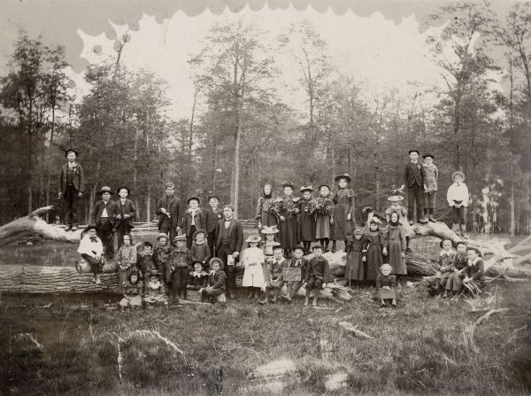 Group of schoolchildren posed sitting and standing on a fallen tree trunk. They are dressed in their finest clothes for a school photograph. One of the boys is holding a small chalkboard on which is written: "The Groves were God's first temples."