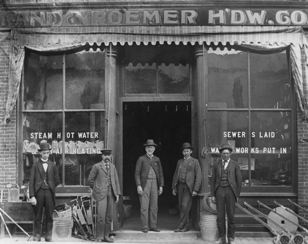 Group of five men standing in front of Rand and Roemer Hardware Company store. Many items can be seen on the sidewalk and through the store windows, including lawnmowers, watering cans, coffee pots, teakettles, a whetstone, washbasins, and bulb planters (?).