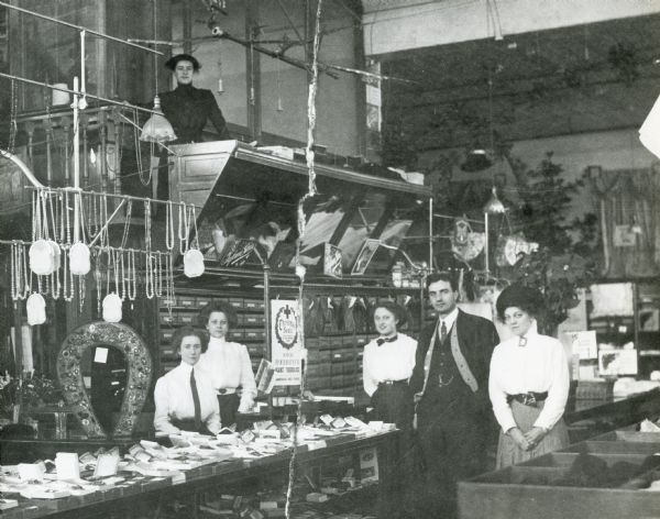 Five female employees and one male employee posed at the jewelry counter of Schuette Brothers Store. There is an American Red Cross Christmas Seals poster displayed behind the counter.