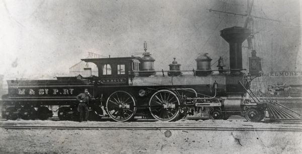 Milwaukee and St. Paul Railway locomotive no. 40, called the L.B. Rock, at the Chestnut Street yards. The mast of a ship is visible in the background, and a man poses against the coal car. There is a sign for Elmore's on the right.