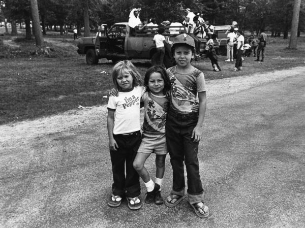 Cuban refugees at Camp McCoy. Three children standing with their arms around each others shoulders. Two wear Woody Woodpecker t-shirts, the other wears a Dr. Pepper t-shirt. In the background, a group of adults on the back of a pickup truck talk and play music. One man has a guitar.