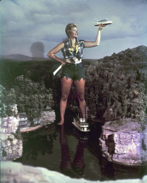 Woman dressed as Paulina Bunyan (female version of Paul Bunyan) standing in the Minirama recreation of the Wisconsin Dells. She holds an axe in one hand and hold a miniature boat up with the other. There is a miniature excursion steamer that runs on tracks between her feet.