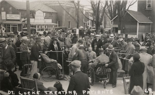 Dr. Mahlon William Locke treating arthritis sufferers on the street in Williamsburg, Ontario, Canada. Many people have gathered to watch or to receive treatment, some in wheelchairs. A coffee shop, a gift shop, and a news stand, as well as cars lining the street, are visible in the background.