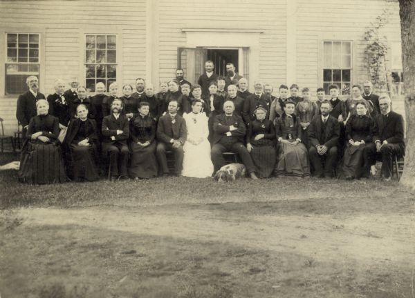 Outdoor group portrait in front of a building of the Wheeler and Wood families. There is a dog laying at the feet of a man seated in the front row.