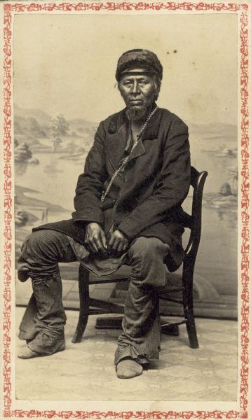 Carte-de-visite studio portrait of man posing on chair in front of a painted backdrop. He has a beard and is wearing a hat, jacket, and trousers.