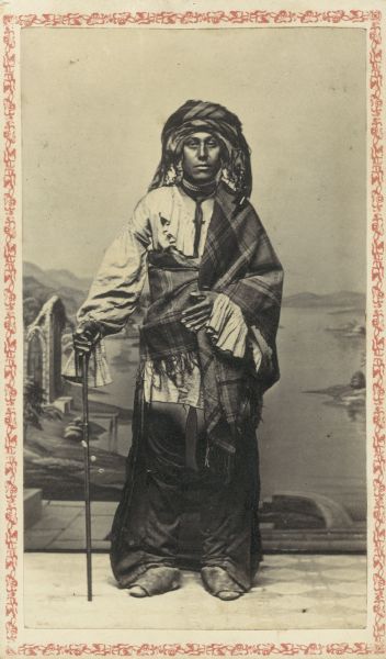 Carte-de-visite studio portrait of man standing with a frilled-bodice, cloth hunting shirt with leather leggings. A wool plaid shawl is worn toga-style; and another is wrapped his head as a turban. He is posing in front of a painted backdrop. Presumably, this young man was son of the Potawatomi Chief Black Hawk, although his moccasins are made in the Winnebago style.