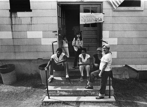 Cuban Family sitting on porch steps with sign on door that says: "Independence 4 of July USA." Photographs made on July 4, 1980, by Archibald of Cuban refugees who had arrived as a result of the Port of Mariel exodus, and were housed at Camp McCoy, Wisconsin.