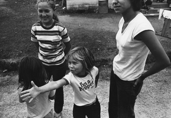 A child extends her hand while standing with other children and an adult. She is wearing a t-shirt with the slogan "I'm a Pepper". Photograph is part of a group of images taken of Cuban refugees who had arrived as a result of the Port of Mariel exodus, and were housed at Camp McCoy, Wisconsin.