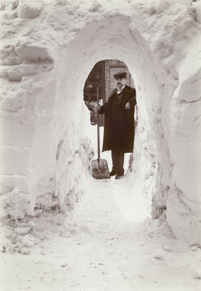 Winter scene of man wearing an overcoat and hat stands with a shovel at the far end of a snow tunnel shoveled through a tall snowbank.