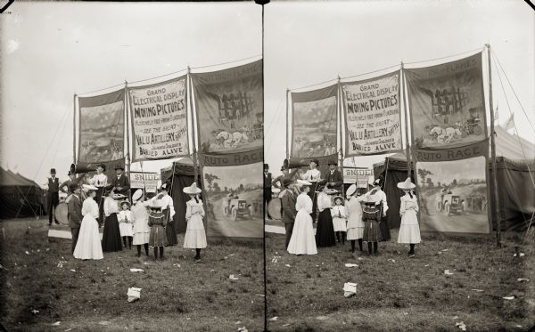 A crowd, including Evaline, Nellie and Harriet Bennett, looking at large banner advertisements for moving pictures. Moving picture subjects include fire fighting and auto races. A man with a megaphone is standing in front of the advertisements and tents.