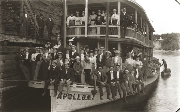 A large group of well-dressed tourists posing on the decks of the steamboat <i>Apollo</i>. A few men are posing on a landing below steep stairs that run along a rock face.