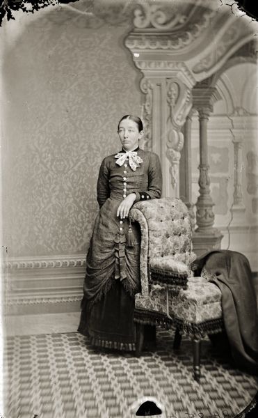 Full-length studio portrait of Francis "Frankie" Irene Douty leaning on a chair in front of a painted backdrop. She was the first wife of Henry Hamilton (H.H.) Bennett.