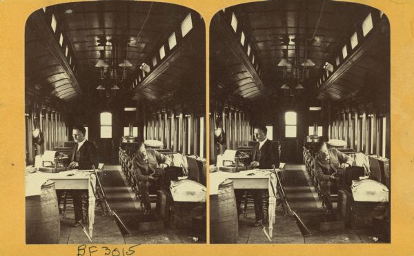 Stereograph of Henry Hamilton Bennett working on a camera in a private car. This is the private car the Wisconsin Central Railroad provided for Bennett and Brainard. Bennett made the exposure by pulling a string attached to the camera. Note the knife and gun he carried with him onboard. In an article published during the trip, he explained they had been hired to portray the beauty along the line. He described Bennett’s “double-barreled” stereo camera and noted the persuasive power of images in the guidebook. Statistics might be fishy, but “photographs never lie.” He is seated near Charles Rollin Brainard, who is working on a typewriter at a desk on the left. There is a rifle propped up between them.