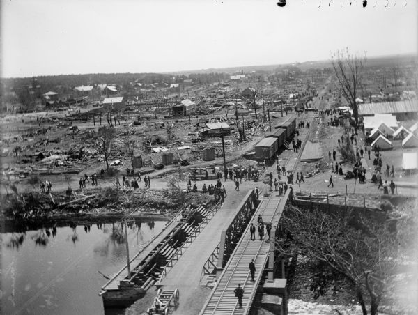Elevated view toward the horizon over a bridge on a river of the widespread destruction left in the wake of a tornado. Destroyed houses litter the scene and train cars are visible on the railroad tracks. Groups of people are scattered throughout the town, and a set of tents are on the bottom right.