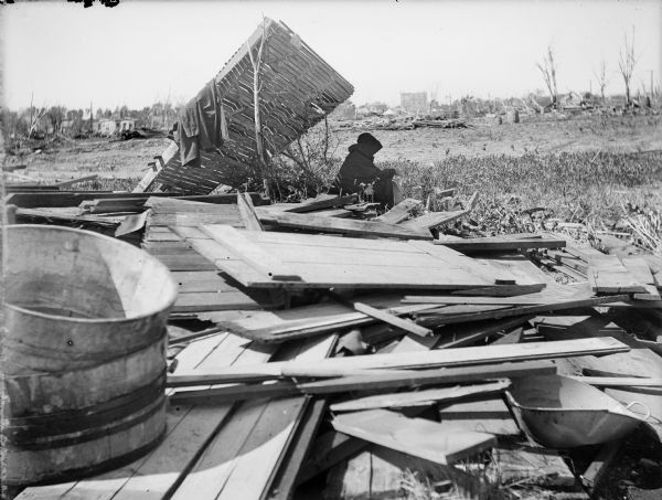 A hooded woman sitting on the remnants of a house. As the amount of debris and barren landscape can attest, the area around her had been leveled by a tornado.