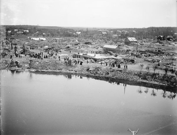 Elevated view over water, perhaps Hatfield Lake, of damage to houses and property caused by a tornado. Groups of people are scattered throughout the town.