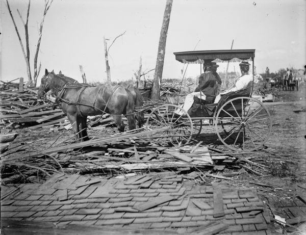 Side view of a group of people looking at the debris left by a tornado from a covered horse-drawn carriage. In the foreground the roof of a houselies on the ground.
