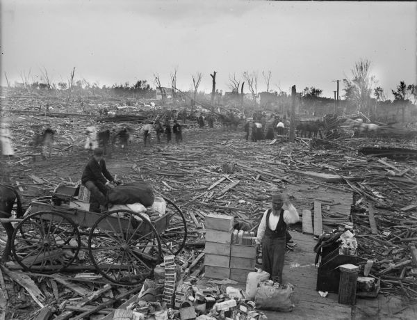 Elevated view of people working with the relief effort in the aftermath of a tornado. Boxes of supplies are piled near a man in the foreground, and a man holds a bundle in a cart parked on the left. In the background people are walking among the debris strewn on the ground.