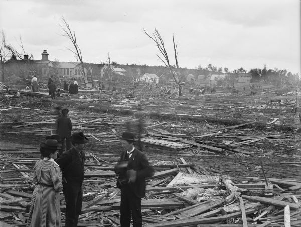 In the foreground a small group of men and women stand surrounded by the debres from a tornado. Storm damaged houses and relief workers are visible in the background. Large buildings in the background on the left appear to be intact.
