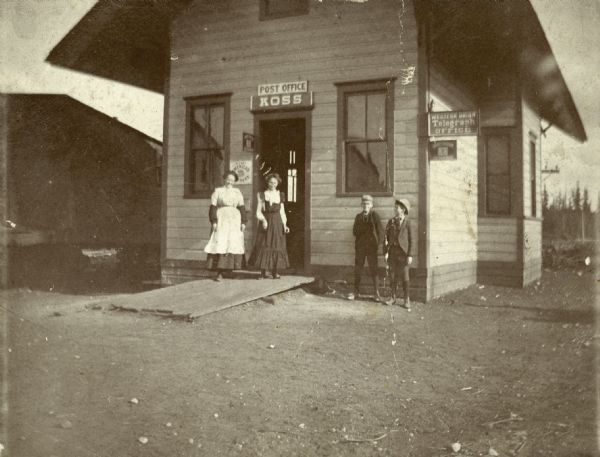 Two women and two young boys pose in front of the Koss Depot, which also serves as a post office and telegraph office.
